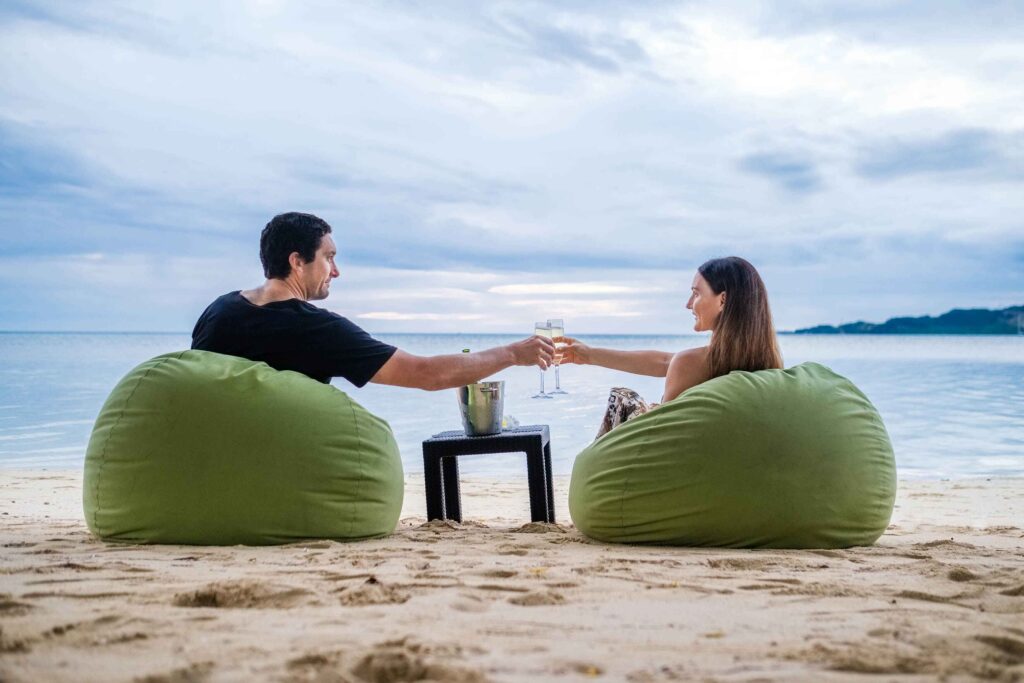 Lomani Island Resort Fiji romantic couple sitting on bean bags on the beach shore drinking champagne