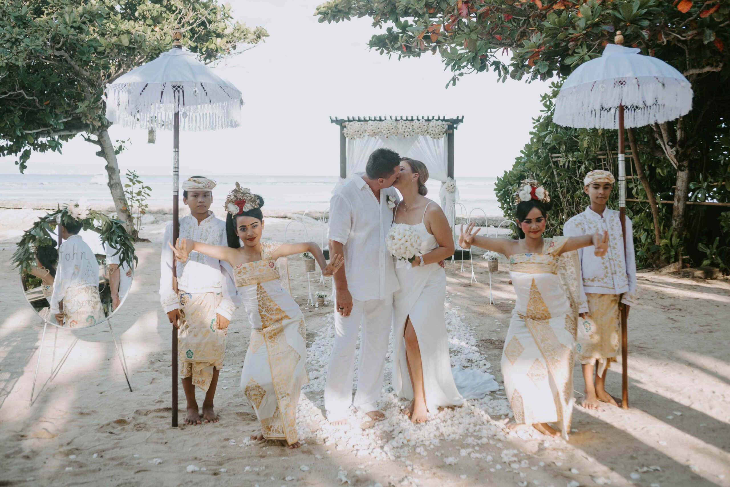 Real wedding John and Teodora at Kayumanis Nusa Dua Private Villa and Spa Bali, Indonesia - post-ceremony-portrait-of-the-bride-and-groom-kissing-with-the-altar-and-crowd-on-the-background
